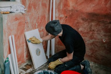 Siyah üniformalı ve sarı eldivenli genç bir beyaz adam dizlerinin üzerinde oturur ve inşaat çalışmalarının devam ettiği bir evde plastik pencere çerçevesinin bir kısmını süngerle yıkar, seçici bir odak ile yakın bir açıdan bakar. Konsept