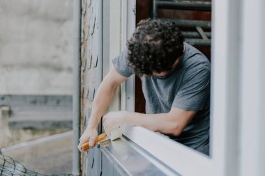 Beyaz, gri üniformalı bir kıvırcık adam pencere çerçevelerini bant ve inşaat bıçağıyla onarıyor. Bir bahar günü odanın içinde dikiliyor. Pencere çerçevesi tamiri, inşaat işi.