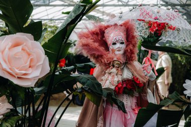 Grand Bigard Şatosu, Belçika - 27 Nisan 2025. Maya çiçek festivaline adanmış bir sergi sırasında, tüylü pembe Venedik kostümlü bir kadın portresi, bir bebek ve bir açılış şemsiyesi kapalı alanda bitkilerin arasında duruyor..