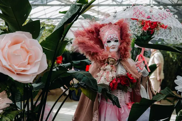 Grand Bigard Şatosu, Belçika - 27 Nisan 2025. Maya çiçek festivaline adanmış bir sergi sırasında, tüylü pembe Venedik kostümlü bir kadın portresi, bir bebek ve bir açılış şemsiyesi kapalı alanda bitkilerin arasında duruyor..