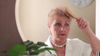 Beyaz bluzlu güzel beyaz yaşlı bir kadın bahar günü yatak odasındaki aynaya ahşap bir tarakla saçını tarıyor. Yan görüntüsü yavaş çekimle çok yakın. Emeklilik yaşam tarzı kavramı.