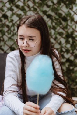 Beyaz tenli güzel bir kızın portresi, gözlüklü, bahçe sandalyesinde oturan, düşünceli bir şekilde kafasını çeviren ve elinde açık mavi bir pamuk şeker tutan, bir bahar güneşli bir günde, arka bahçesinde elinde bir çubuk tutan,