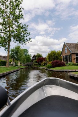 Güzel dikey kırsal manzara, bulutlu bir bahar gününde dar kanaldaki evler arasında tahta köprülere doğru giden metal gri tekne kıçı, yan görüş planı. Yat zamanı, seyahat zamanı, lüks hayat, doğa geçmişi..