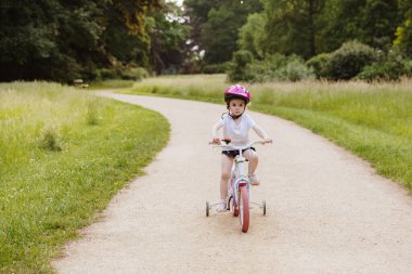 Parlak pembe kasklı, güzel, beyaz bir kızın portresi. Pembe ve beyaz bisiklet sürüyor. Bahar günü parkta tek başına. Mutlu çocukluk kavramı, bisiklete binen çocuklar.