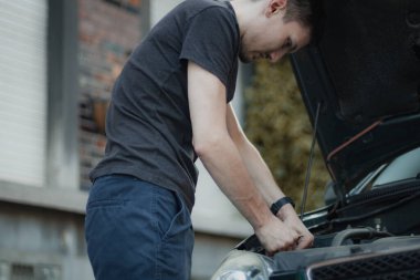 Genç beyaz bir adam bir arabanın ön farında lambayı söküyor, bir yaz öğleden sonra şehrin bir sokağında yarım dönüş yapıyor, aşağıdan yakın çekim manzarası var. Ampulleri ve araba tamirini değiştirme kavramı.