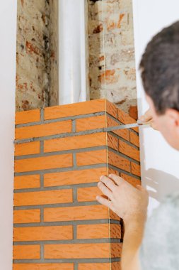 Genç bir beyaz adam, bir eliyle şömine bacasındaki tuğlaların arasında dar ve uzun bir spatula kullanarak aşırı taze çimento harcı ile temizlik yapar, gün boyunca bir sandalyenin üzerinde dururken, yan görüntüyü seçici bir odak ile yakın plan yapar. Temiz