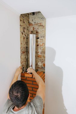 Genç beyaz bir adam arkada duruyor ve taze çimento harcı düzleştiriyor, bir tuğlayı çekiçle vuruyor, şöminenin bacasında temiz bir tuğla yapıyor, yan görünüş seçici bir odak ile kapanıyor. Temiz tuğla işi kavramı, aile evi