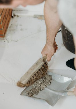 Genç bir beyaz erkek müteahhit, bir süpürgeyle uzanan tuğlanın ortasında kurumuş çimento kalıntılarını süpürüyor ve odanın içinde bükülmüş vaziyette faraş gibi duruyor. Yan görünüş yakın. Temiz tuğla işi konsepti, ev yenilemesi..