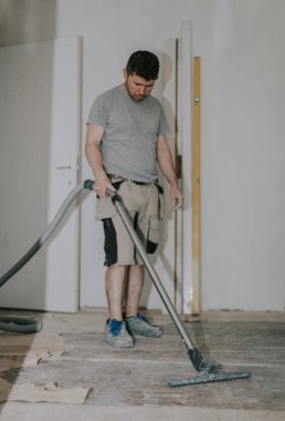 Young caucasian brunette man in work clothes standing in front vacuums the floor with a construction vacuum cleaner in a room in an old house where renovations are taking place, bottom view close-up with selective focus.Construction work concept.