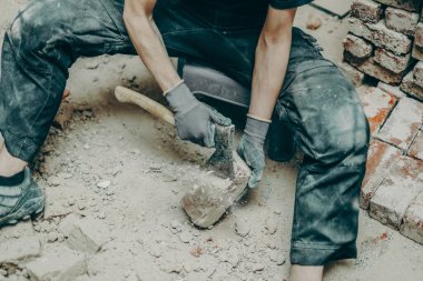 Siyah iş kıyafetleri içinde tanınmayan genç, beyaz bir adam, küçük bir sandalyede oturuyor, bacakları açık, toprak ve eski çimentodan tuğlaları temizliyor, balta kullanıyor, üst tarafı yakın plan. İnşaat işi kavramı.