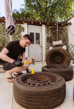 Dikey portrede, çekiç ve sarı-siyah levye kullanan, eski lastikleri direksiyondan söken, bahar günü arka bahçede çömelmiş, yan görüş planı yapan, gözlüklü yakışıklı bir genç var. Araba lastiklerini çıkarma, tamir etme kavramı