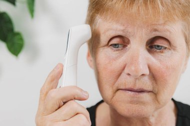 Yaşlı, güzel bir kadının portresi, gri gözlü ve sıcaklığı ölçmek için tapınağın yakınındaki termometreyi kullanmayan, gündüz vakti odada, çok yakından bakıyor. Teknoloji ve emeklilik yaşam tarzı kullanma kavramı.