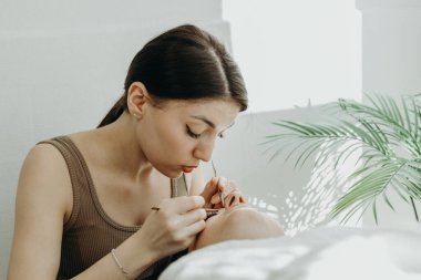 Genç ve güzel bir beyaz kozmetik uzmanı kızın portresi. Bir bahar günü evinde kozmetoloji masasında uzanan kadın müşteriye iki cımbız yardımıyla sağ gözüne yapay kirpikler yapıştırıyor.