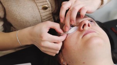 Genç bir beyaz kozmetik uzmanı, bir bahar günü evinde kozmetoloji masasında uzanan bir kadına ellerini kullanarak sağ gözkapağının alt kısmına beyaz bir leke yapıştırıyor. Yavaş çekim ve alan derinliği ile yakın plan görüntüsü.
