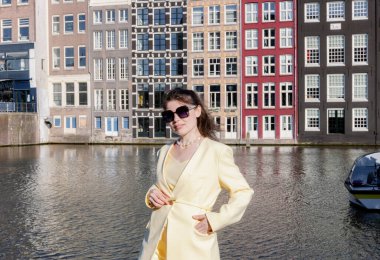 Güneş gözlüklü ve sarı tereyağlı güzel beyaz bir kızın kalça boyunda portresi. Güneşli bahar günlerinde, kentteki renkli evlerde kameraya bakıp gülümsüyor. Şehir içi yürüyüşü, yaşam barınağı