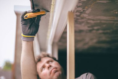 Gri eldivenli, genç, kıvırcık saçlı beyaz bir adam pencerenin dışından bakan, iskelede duruyor, sarı bir inşaat bıçağıyla pencereyi temizliyor, evin ikinci katında yenileme için hazırlıyor,