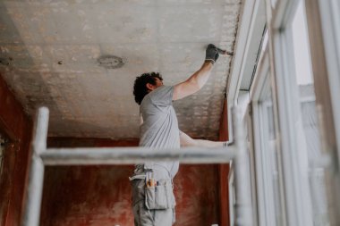 Genç bir Kafkasyalı iskelenin yanlamasına dikiliyor ve yenileme çalışmalarının devam ettiği bir evin tavanı olan pencerenin çatısındaki eski montaj köpüğünü temizlemek için bir yapboz kullanıyor. Hom