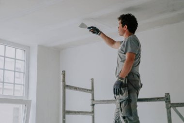 Üniformalı yakışıklı beyaz bir adam inşaat ve yenileme yapılan bir odanın tavanını spatulayla temizliyor. Alt tarafı derin bir manzara. Ev yenileme ve tavan temizliği.