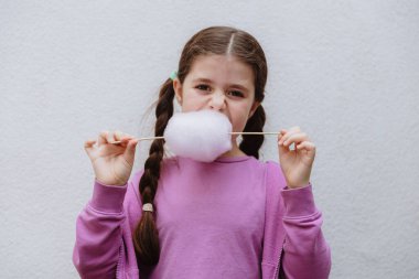 Esmer, güzel bir kızın belden yukarı portresi, leylak rengi elbiseli, bir çubuk üzerinde hafif pamuk şekeri ısıran, güneşli bir bahar gününde arka bahçede beyaz bir duvarın yanında duran, kenar manzaralı. Pamuk helva zamanı.