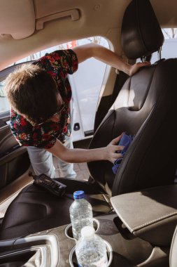 Beyaz tenli genç bir esmer adamın portresi. Güneşli bir bahar gününde araba yıkama yerinde ön yolcu koltuğunu mavi bez ile temizlerken orta ölçekli bir fotoğraf. Konsept araba yıkama, çalışan insanlar.