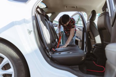 Güneşli bir bahar gününde şehrin araba yıkama yerinin solundan arabanın arka yolcu koltuğunu süpüren beyaz, yakışıklı bir adamın portresi. Yan taraftan manzara, yakın çekim, orta boy. Kavram: zaman