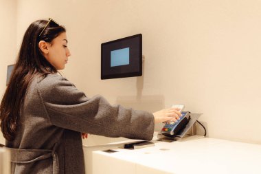 Gri montlu, beyaz tenli genç bir kız, cep telefonunu terminale uygulayarak, alışveriş merkezindeki bir mağazada dikerek, kendi başına ödeme yapıyor. Çevrimdışı alışveriş konsepti, moda yaşam tarzı, alışveriş