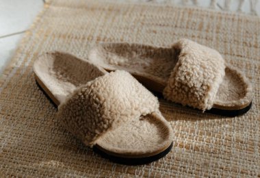 Güneşli bir günde, beyaz fayanslı bir odada, kahverengi renkli koyun derisi ve mantar tabanlı bir çift güzel ev modası, şık ve sıcak terlik, jüt bir kilimin üzerinde duruyor. Ev ayakkabısı konsepti.