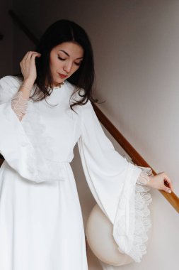 Beyaz uzun elbiseli, beyaz saçlı, beyaz saçlı güzel bir esmer kızın dizlerine portre, elleriyle saçlarını ayarlıyor, bir bahar günü, evin ahşap merdivenlerinden dikkatlice iniyor, yakın çekim. Yaşam tarzını kavra