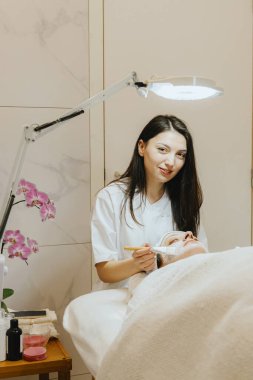 Genç, beyaz tenli kozmetik uzmanı kameraya gülümseyerek bakar ve beyaz krem bir maske takar. Burnunun ve dudaklarının çevresindeki yetişkin bir müşterinin yüzüne sürterek masaj masasında gözleri kapalı bir şekilde uzanır.