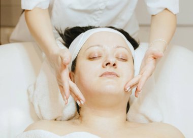 Tanınmayan genç bir kozmetik uzmanı parmaklarıyla boynuna ve çenesine nazikçe masaj yapıyor. Yetişkin bir müşteriye, gözlerini kapalı bir masaj masasına yatırıyor..