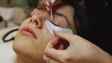 Beyaz tenli genç bir kozmetik uzmanı pamuk çubuğuyla göz kapaklarını temizliyor ve genç bir müşteriye sıvı solüsyon veriyor. Bir güzellik salonunda yatıyor. Kirpik kaldırma konsepti, güzellik endüstrisi..