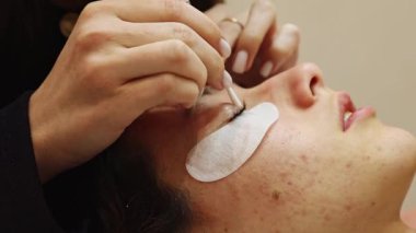 Sağ gözünün kapağından tutkal temizleyen genç bir beyaz kız kozmetik uzmanı genç bir müşteriye pamuk çubuğuyla uzanan genç bir müşteri bir güzellik salonunda yatıyor. Saha derinliği ile yakın plan çekimde. Kirpik kaldırma konsepti, güzellik endüstrisi..