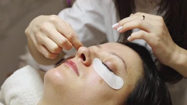 Kaplamasını bitirdikten sonra sol gözünden yapışkan bir yamayı çıkaran genç bir beyaz kız kozmetik uzmanı. Bir müşteri, bir güzellik salonunda kozmetoloji masasında yatıyor. Kirpik kaplama konsepti, güzellik endüstrisi..