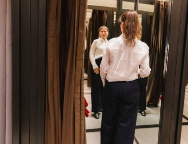 Arkadan güzel, beyaz, olgun bir kızın portresi, soyunma odasında duruyor ve bir bahar günü alışveriş merkezindeki bir butiğin aynasında yeni kot kıyafetini inceliyor, yan görünüm, yakın çekim. Konsept, alışveriş zamanı