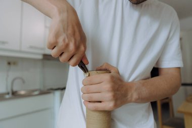 Beyaz tenli genç bir adam tamir etmek için kedi evinin karton bir tübündeki penseyle zımba tellerini söker. Yazın mutfakta durur. Arka planda bulanıklık vardır. Bir kediyi tamir etme kavramı