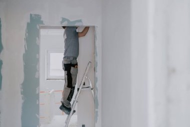 Gri tişörtlü genç bir beyaz adam, bir eliyle bir baltayla kapı aralığındaki eski macunu temizliyor. Bir yandan merdivenin kenarında dikilirken, diğer yandan seçici bir odak noktasıyla. İnşaat işi kavramı.