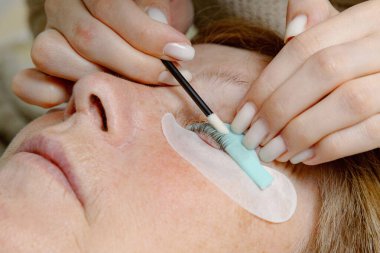 Beyaz tenli genç bir kozmetik uzmanı sol gözünün kirpiklerini sünger çubuğuyla düzeltiyor ve onları bir güzellik salonundaki kozmetik yatağında uzanan yaşlı bir kadın müşterinin silikonuna çekiyor.