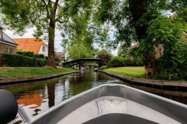 Bulutlu bir bahar gününde, dar kanalda tahta köprülere doğru yelken açan, metal gri tekne kıçı manzaralı güzel bir kır manzarası. Yat zamanı, seyahat zamanı, lüks hayat, doğa geçmişi..