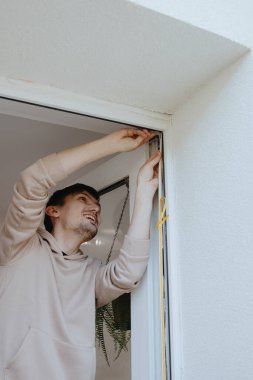 Kafkasyalı mutlu bir gencin portresi. Kapı çerçevelerinin üst köşesine kendi kendine yapıştırıcı profil yapıştırıyor. Sandalyenin üstünde duruyor. Pencere çerçevelerinin tamiri ve yalıtımı kavramı, bahar çalışması.