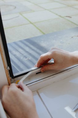 Güneşli bir bahar gününde, plastik pencere çerçevesinin sol alt köşesine, çok yakından görünen beyaz, içi boş, kendi yapıştırıcı kauçuk bir profil yapıştıran genç bir beyaz adamın portresi. Tamir, yıkama ve yalıtım konsepti