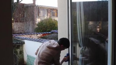 Genç, beyaz bir adam pencerenin çerçevesini mavi bir mikrofiber bez kullanarak yıkıyor, bardağa yansıyor ve ikinci kattaki bir odada dikiliyor, yan görüntü orta ölçekli ve yavaş çekte çekiliyor. Pencerenin onarımı ve yalıtımı kavramı