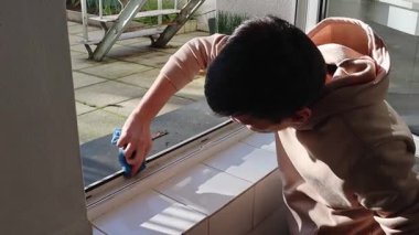 Yakışıklı beyaz adam, kirli iç pencere çerçevelerini mavi mikrofiber kumaşla yıkıyor kauçuk profili yapıştırmadan önce, yan görüntüyü yavaş çekimle çekiyor. Pencere çerçevelerinin onarımı, yıkanması ve yalıtımı, ilkbahar işi.