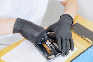 Beyaz tişörtlü, siyah lateks eldivenli, tanınmayan genç bir beyaz adam toz ve tozdan akıllı bir telefonun arkasını temizliyor, yaz günü yakın çekimde, mutfakta bir masada oturuyor. Cep telefonu tamir konsepti, yeni hayat.