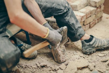 A young caucasian man in black work clothes sits on a small chair on the left and cleans the bricks from dirt, debris and old cement with an ax, side view close-up.Construction work concept.