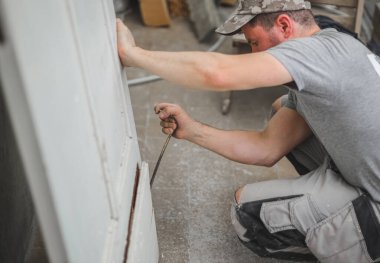 Genç yakışıklı beyaz bir işçinin portresi, eski ahşap bir kapıyı levyeyle söküyor, levyeyi çatlağa itiyor ve onarımların devam ettiği bir odada çömelirken bir tahtayı sıkıyor, yakından bakıyor. Konsept inşaat işi