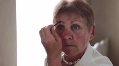 Beyaz bluzlu, beyaz kaşlarını fırçayla boyayan, bahar günü yatak odasındaki aynaya bakan beyaz, yaşlı, güzel bir kadın. Yan görüntüsü yavaş çekimde. Emeklilik yaşam tarzı kavramı.