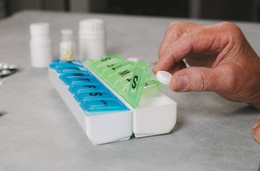Beyaz bir kadının eli plastik organizatörün kompartımanına hap koyar, gri masada oturur, yan görüş çok yakındır. Hapları düzenleyicide depolama kavramı.