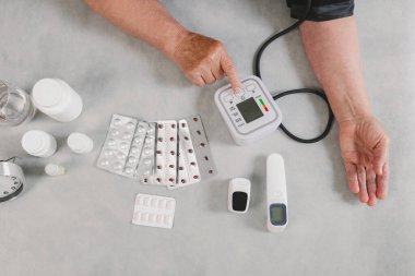 Yaşlı kadının yatay portresi, kan basıncını tonometreyle ölçüyor gri masada oturuyor ve gündüzleri ilaç hazırlanıyor. Üst mercek yakın plan. Teknoloji ve emeklilik yaşam tarzı kullanma kavramı.