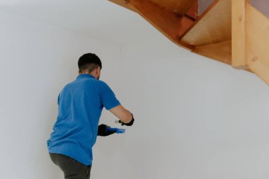 Mavi tişörtlü, yakışıklı, koyu renk saçlı beyaz bir adam arka tarafta duruyor ve fırçayı beyaz boyaya batırıyor, alt tarafı seçici bir odak noktasıyla yakın plan görüşe sahip..