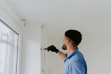 Mavi tişörtlü, koyu renk sakallı, beyaz saçlı genç bir adam pencerenin kenarında duruyor ve bir duvarı beyaz boyayla boyuyor..